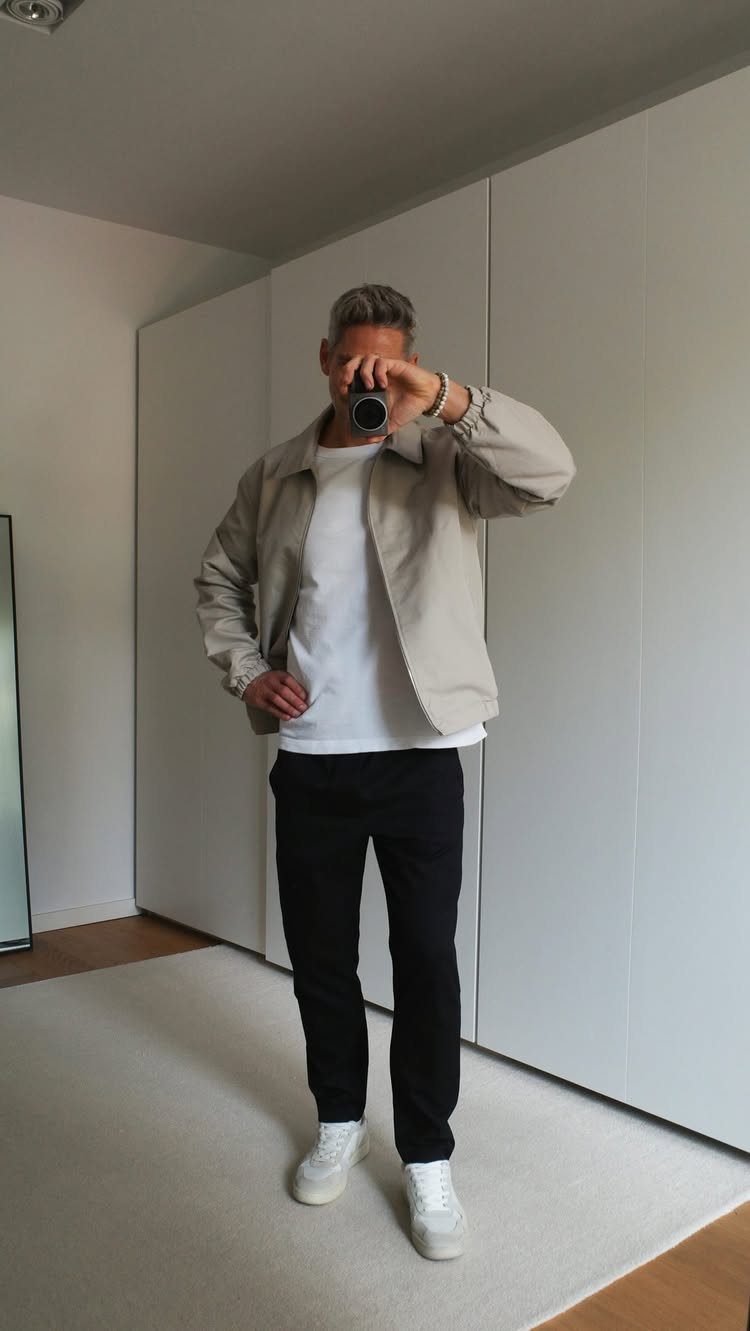 A man posing in a beige jacket, white t-shirt, black pants, and white sneakers.
