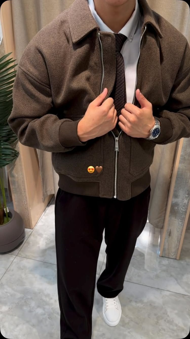Man wearing a brown bomber jacket, white shirt, dark tie, black trousers, and white sneakers.
