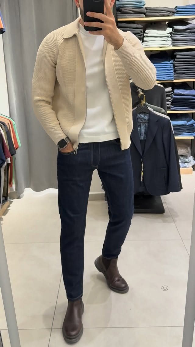 Man in a cream cardigan, white t-shirt, navy jeans, and brown Chelsea boots taking a selfie.