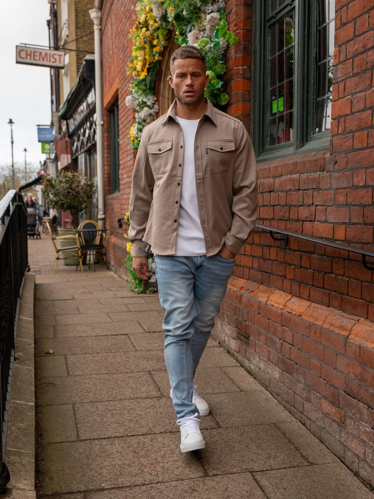 A man in a tan shirt, white t-shirt, and light blue jeans walking outdoors.