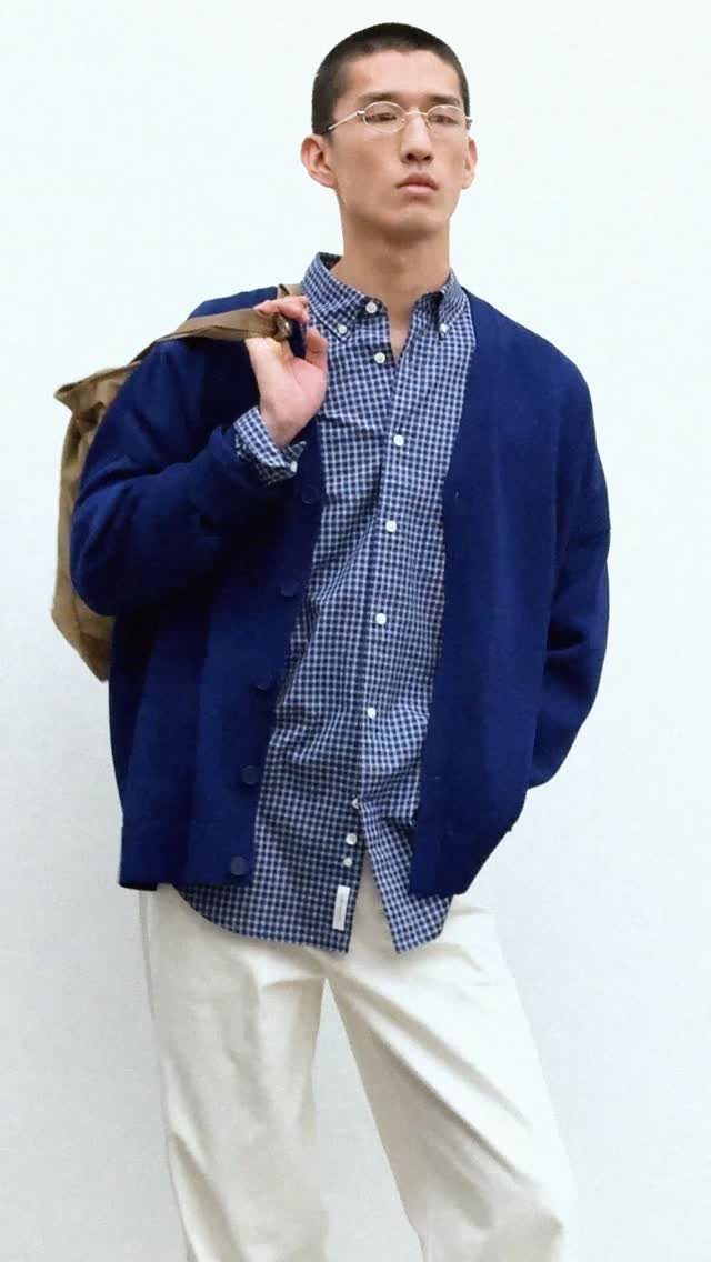 Man wearing a blue checkered shirt, navy cardigan, and white trousers against a white background.