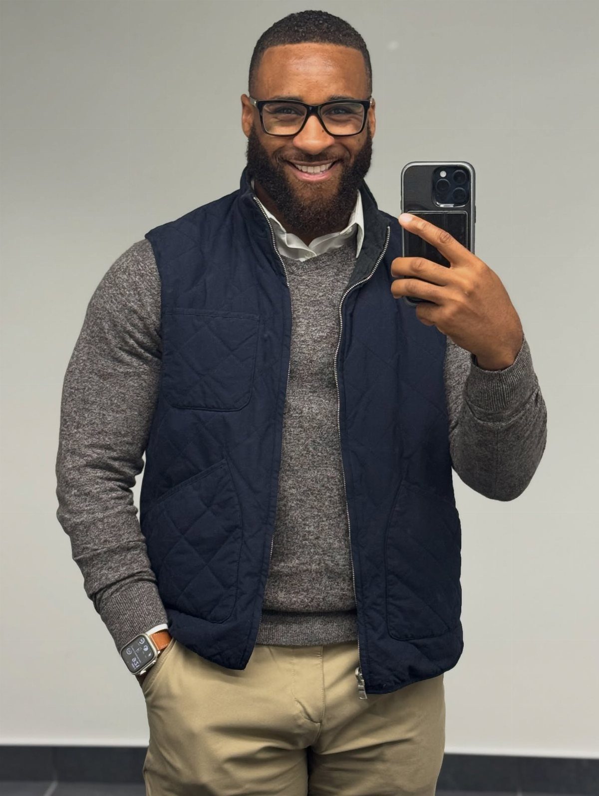 Smiling man in a grey sweater, navy quilted vest, and khaki chinos taking a selfie.