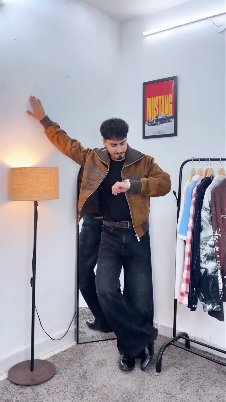 Man wearing a brown suede jacket, black shirt, and dark wide-leg jeans posing indoors.