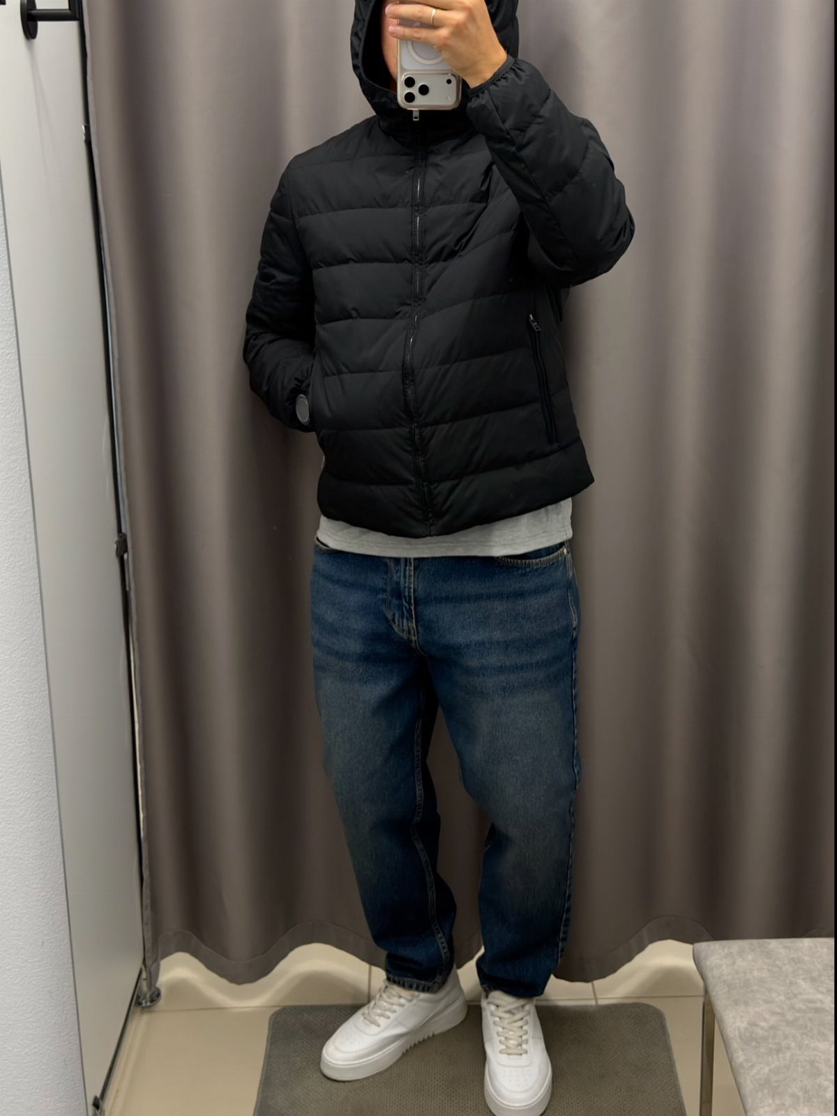 A person wearing a black puffer jacket, blue jeans, and white sneakers standing in a fitting room.