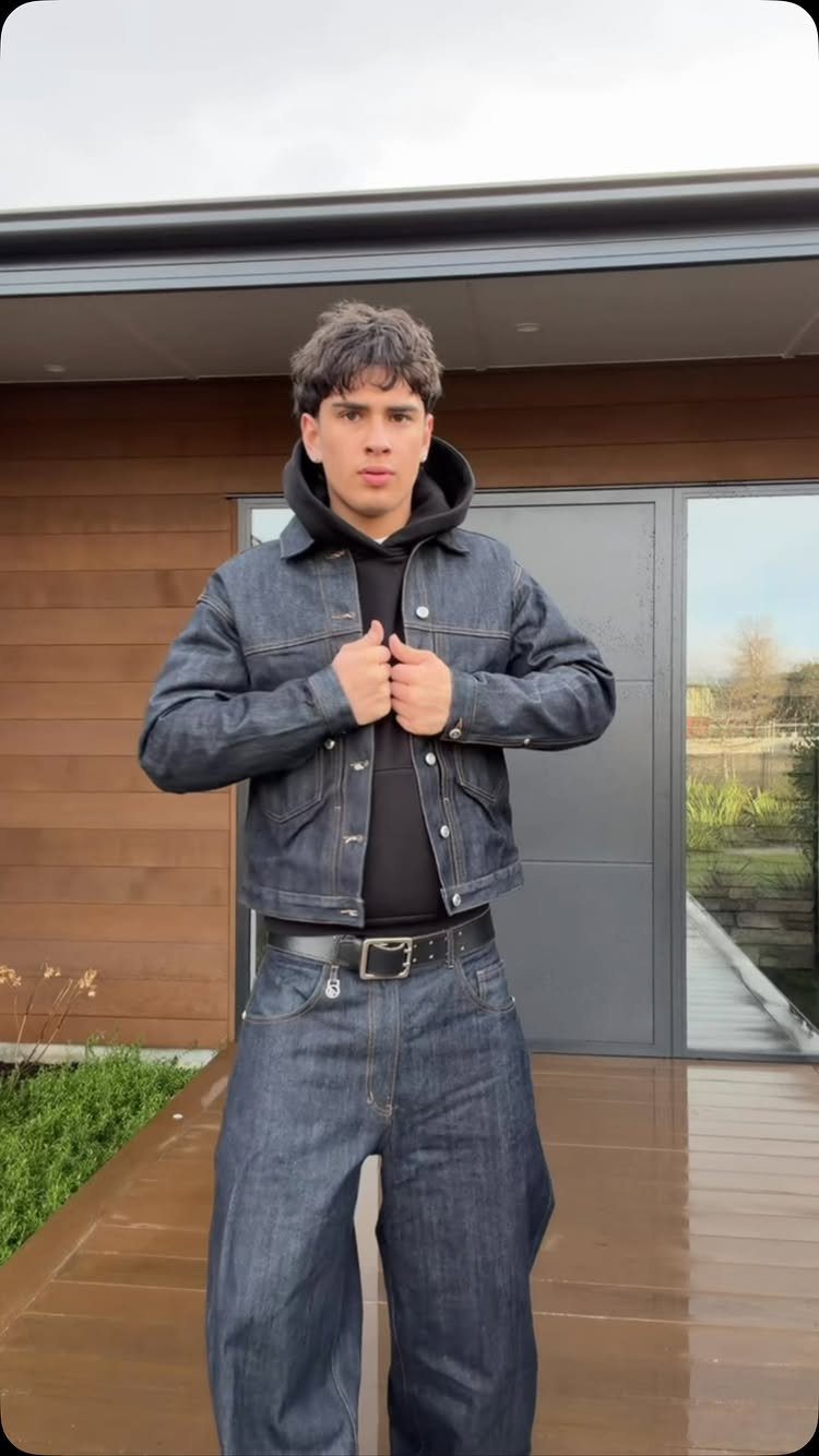 Young man posing in a dark denim jacket, black hoodie, and wide-leg denim jeans.