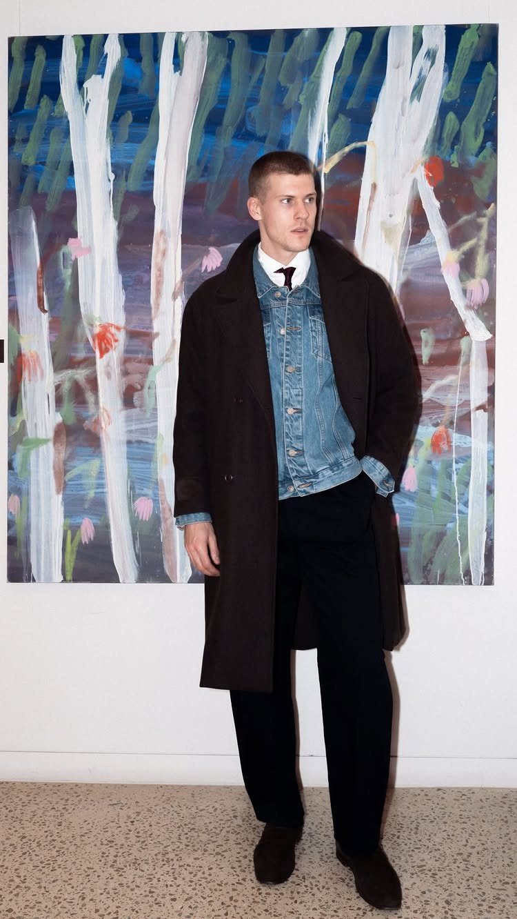 Man wearing a long dark brown coat over a denim jacket and dark trousers.