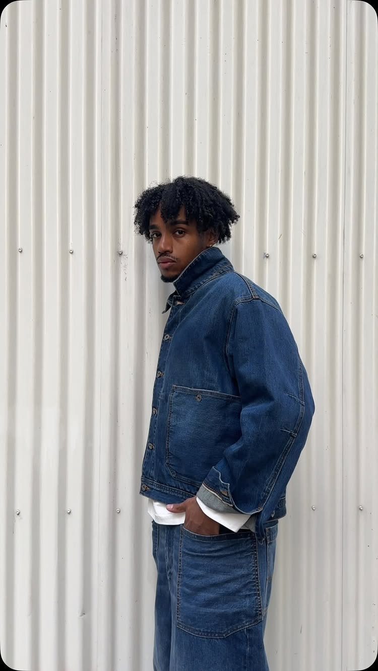 A young man wearing a dark blue denim jacket and matching jeans against a white wall.