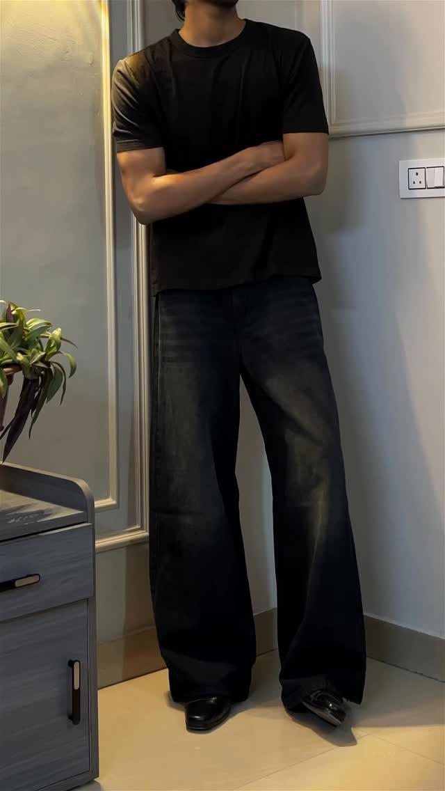 A person wearing a black t-shirt and dark wide-leg jeans standing against a wall.