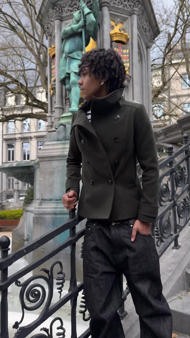 A man wearing a dark olive green pea coat and black jeans standing outdoors.