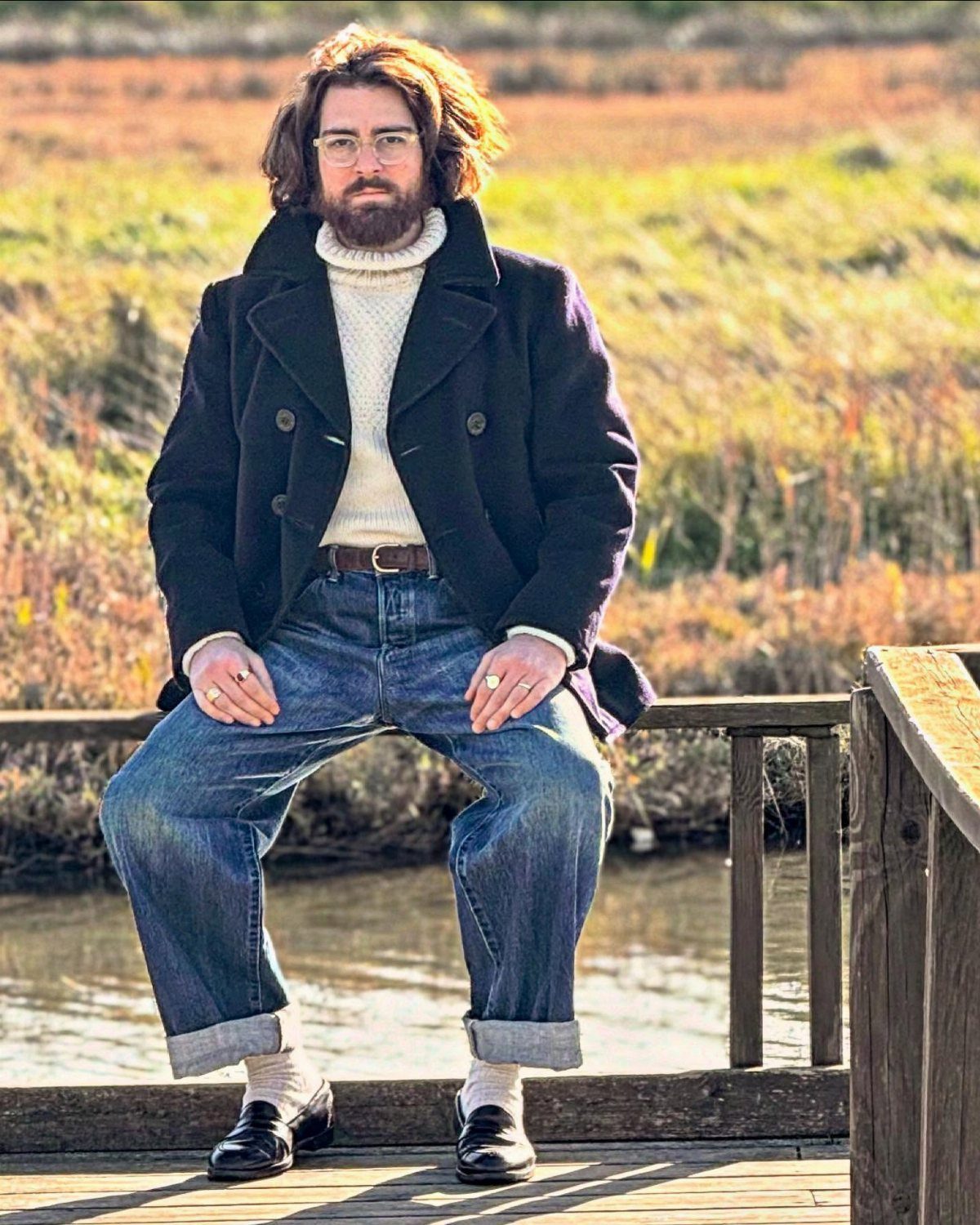 Man wearing a navy pea coat, white turtleneck, and blue denim jeans sitting on a wooden railing.