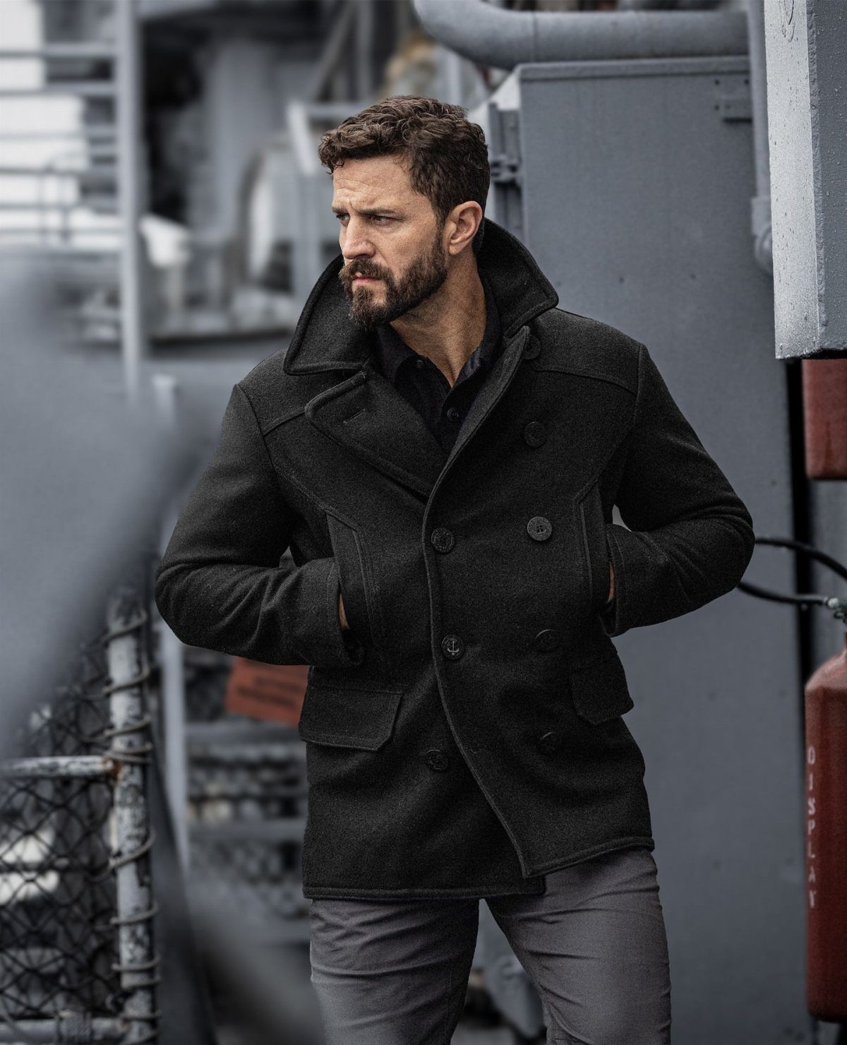 Man wearing a dark charcoal pea coat and grey trousers standing in an industrial setting.