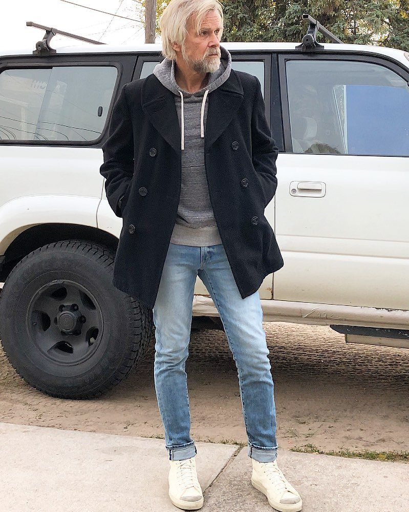 Man with grey hair wearing a black pea coat, grey hoodie, and blue jeans standing by a white SUV.