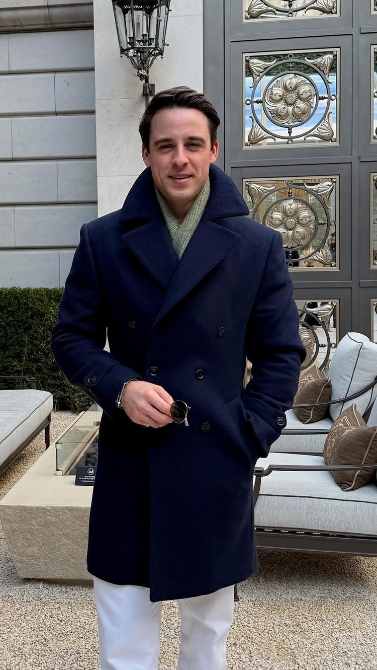 Man wearing a navy blue pea coat, light green scarf, and white trousers standing outdoors.