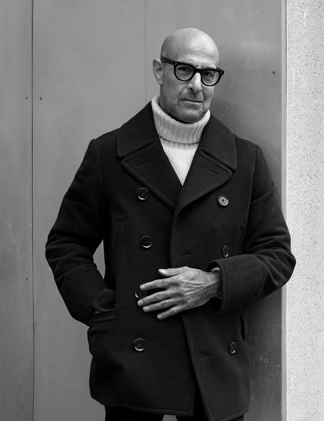 A man wearing a dark pea coat and white turtleneck sweater posing against a wall.
