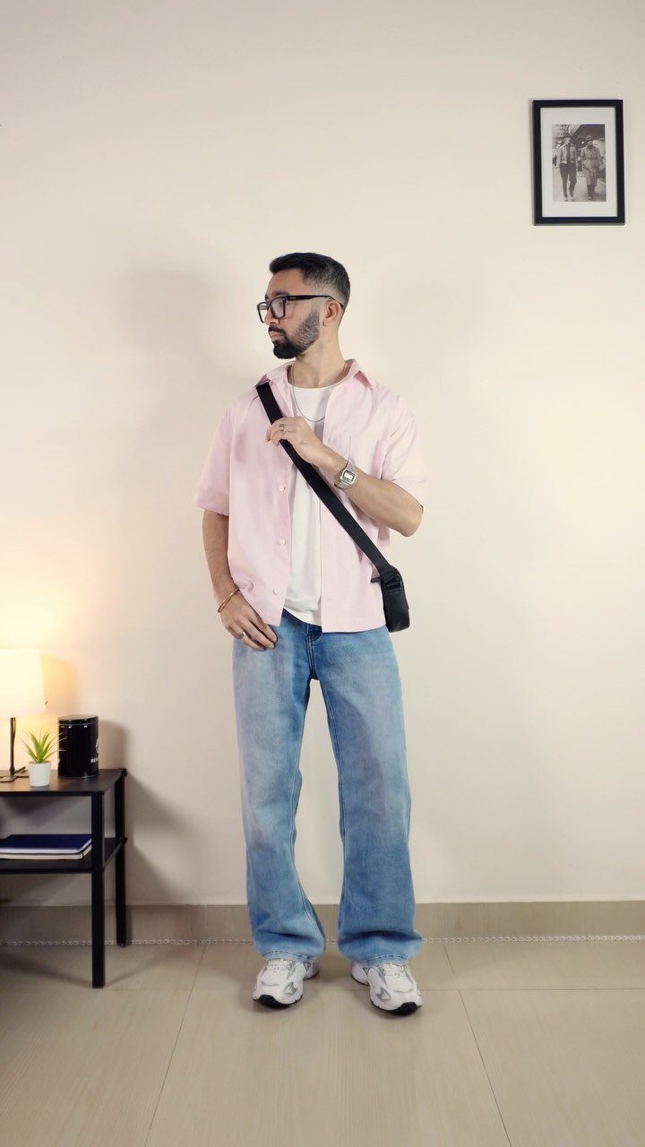 Man in a pink shirt and light blue jeans standing against a neutral wall.