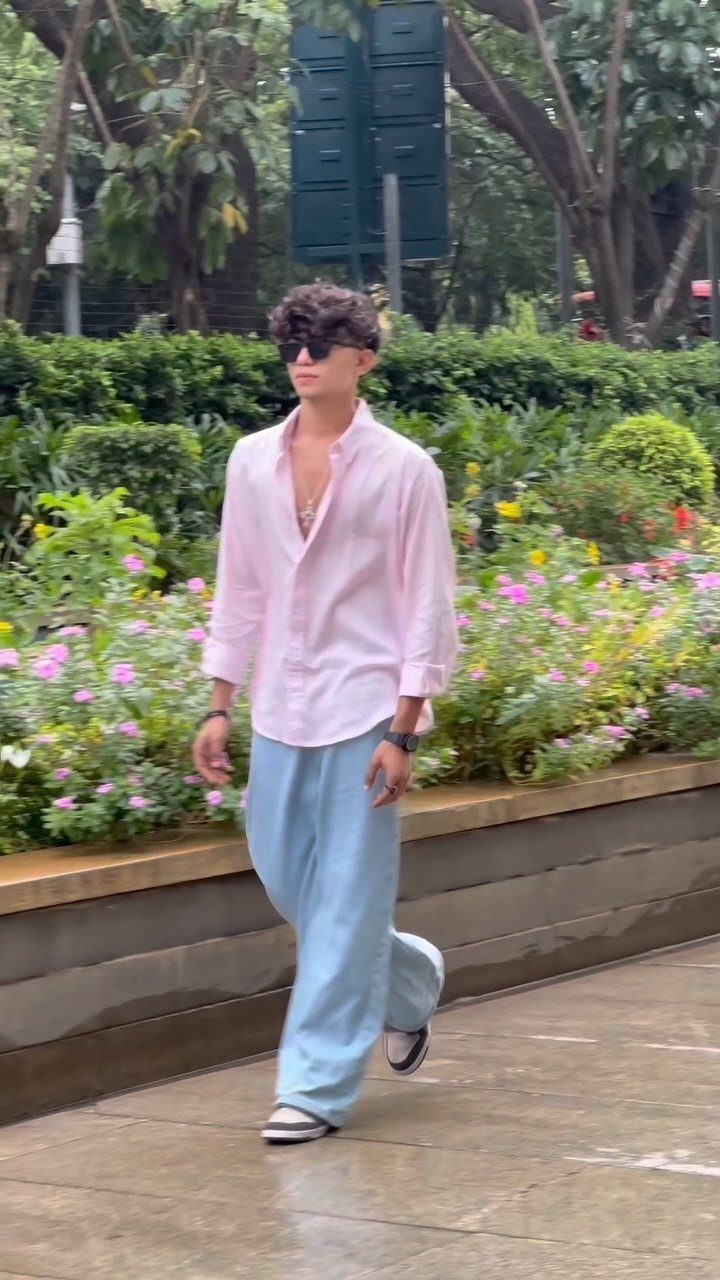 Man in a pink shirt and light blue wide-leg trousers walking outdoors.