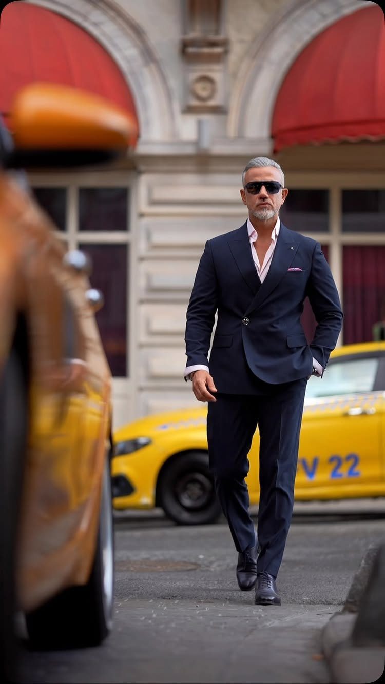 Man in a navy suit and pink shirt walking past a yellow taxi on a city street.