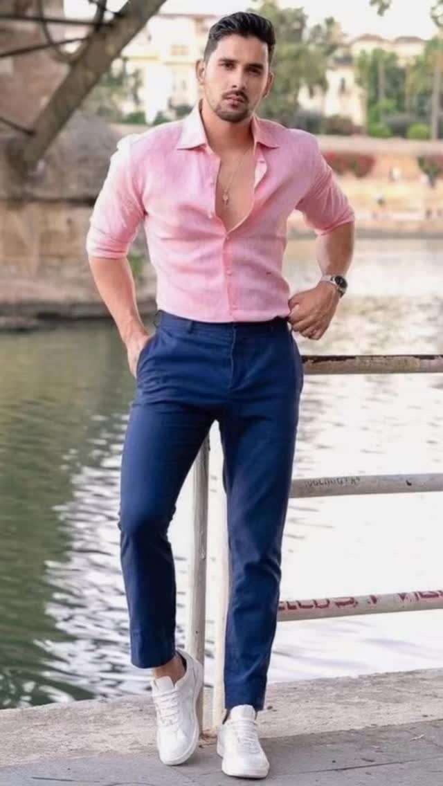 Man in a pink shirt and navy trousers posing by a river.