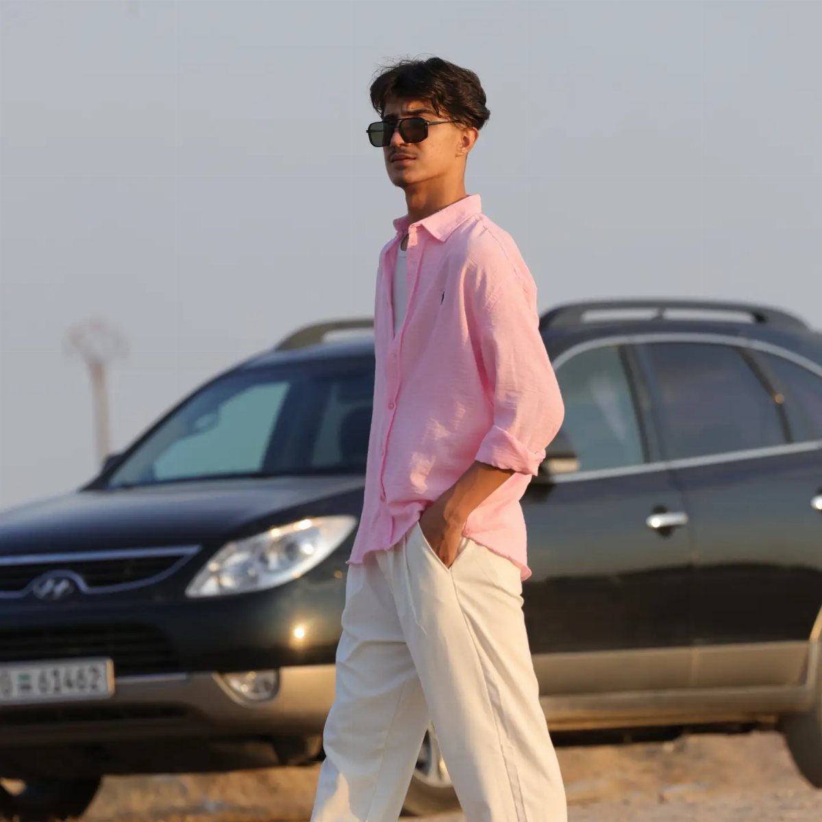 A man in a pink shirt and white trousers walking past a dark car.