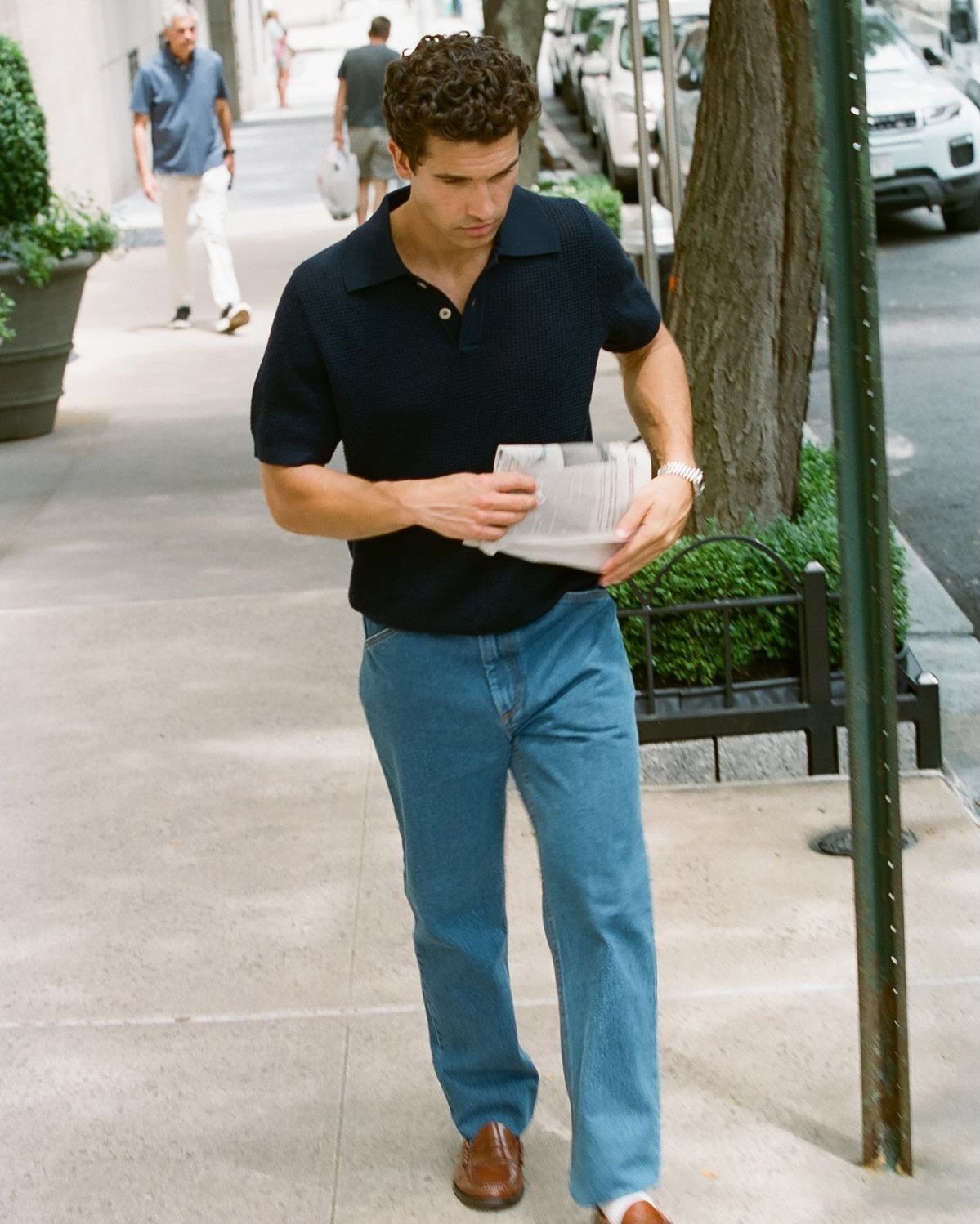 Man in a black polo shirt and blue jeans walking on a city sidewalk while reading a newspaper.