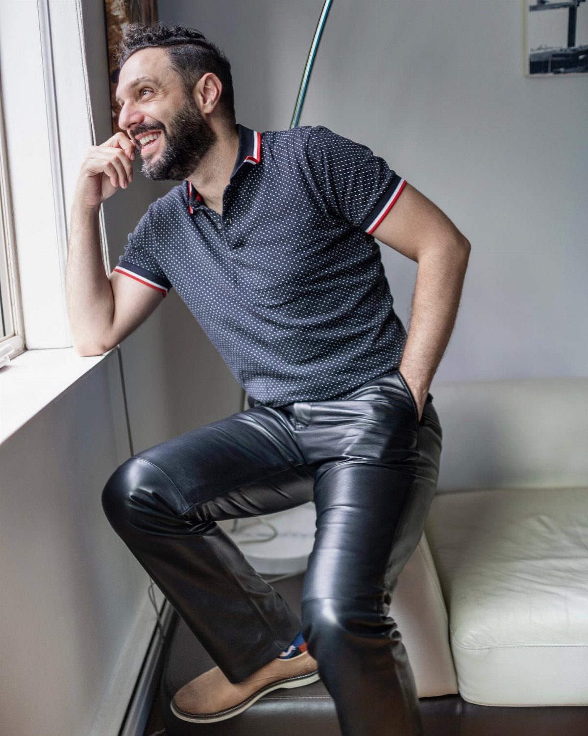 A bearded man in a navy polka-dot polo shirt and black leather pants leans against a window.