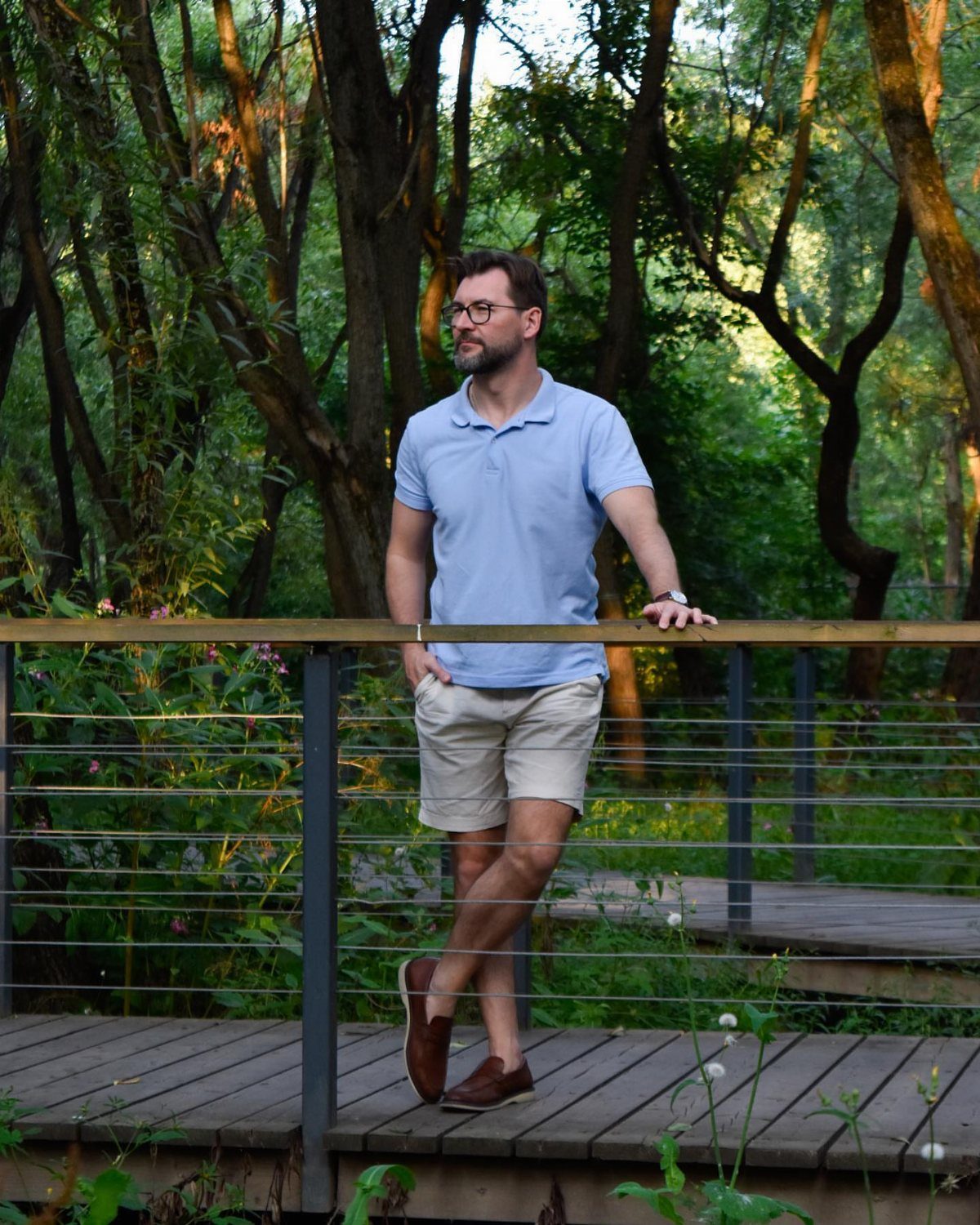 Man in a light blue polo shirt and beige shorts standing on a wooden bridge in the woods.