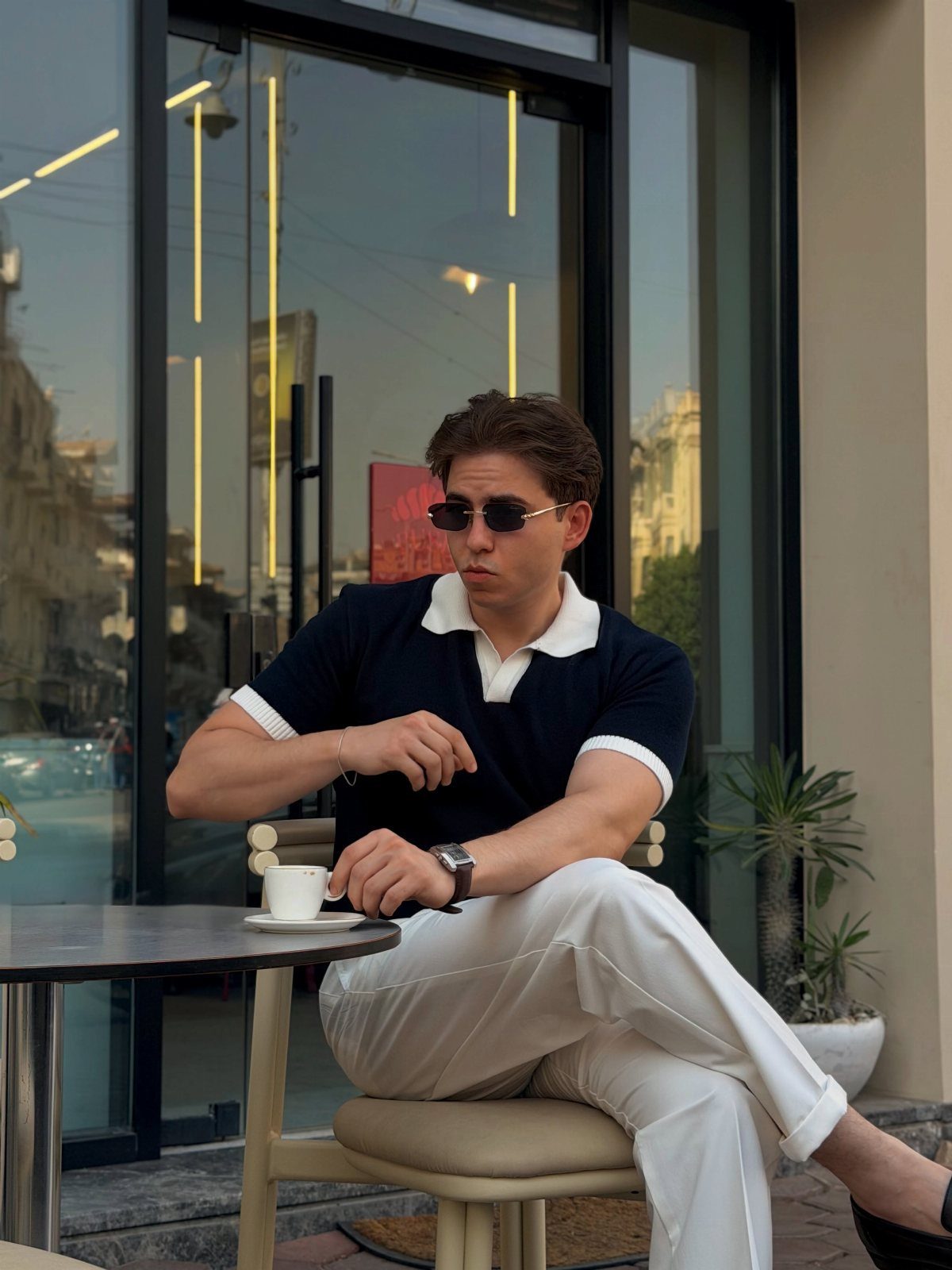 Man in navy polo shirt and white trousers sitting at an outdoor cafe table.