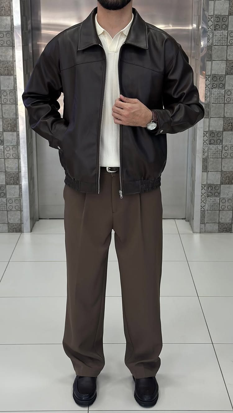 Man wearing a white polo, brown leather jacket, brown trousers, and black loafers standing indoors.