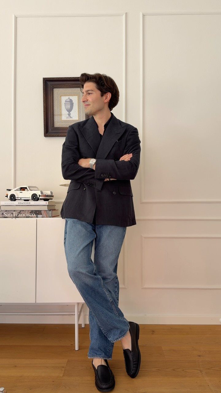 A man in a navy blazer, dark polo, blue jeans, and black loafers standing indoors.
