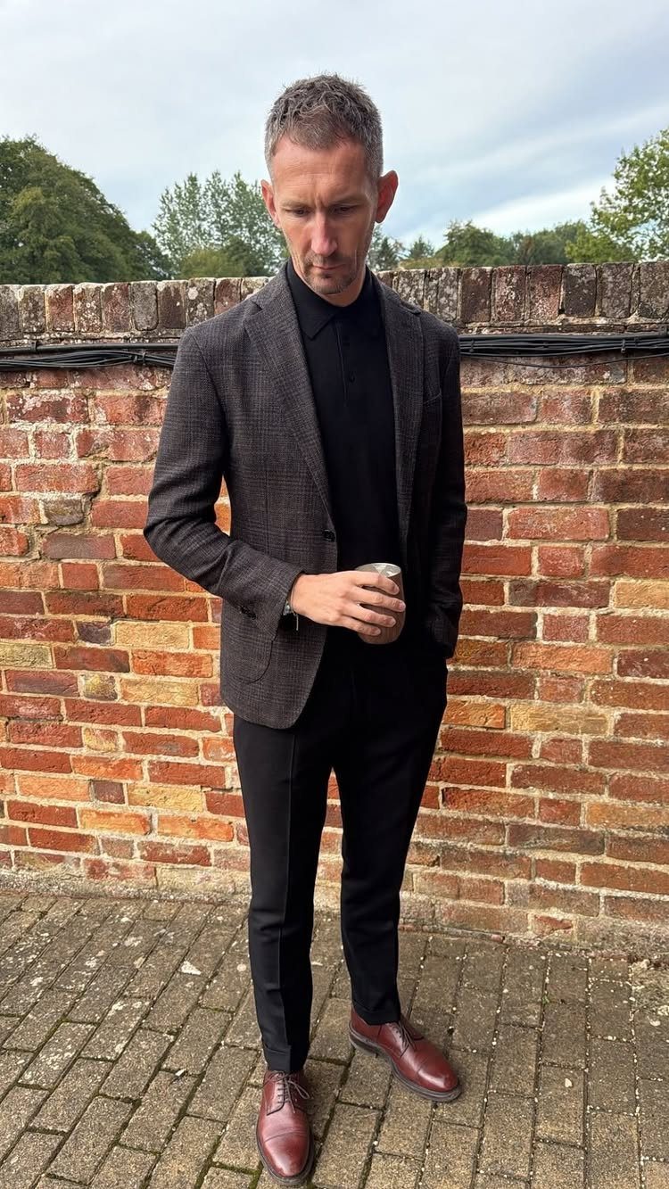 Man wearing a dark patterned blazer, solid black polo shirt, black trousers, and reddish-brown shoes.