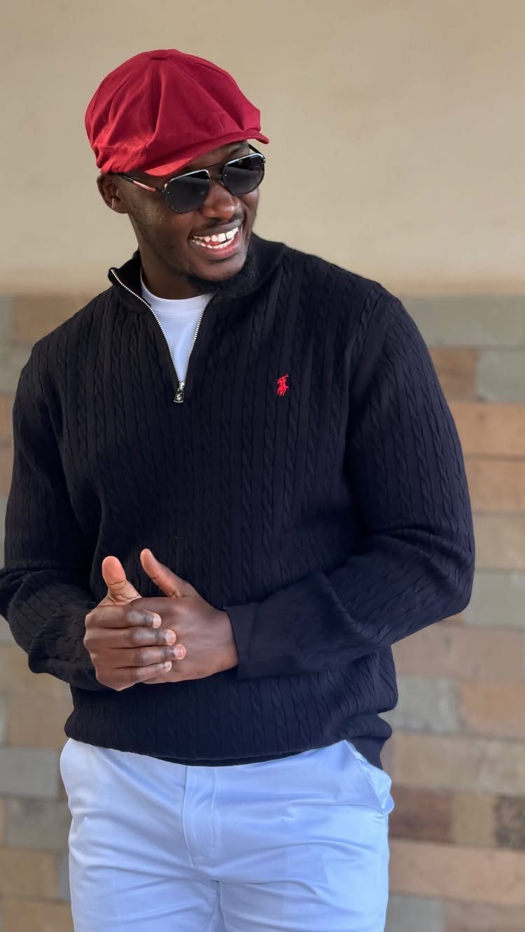 Man smiling in a black textured quarter-zip sweater and white trousers wearing a red beret.