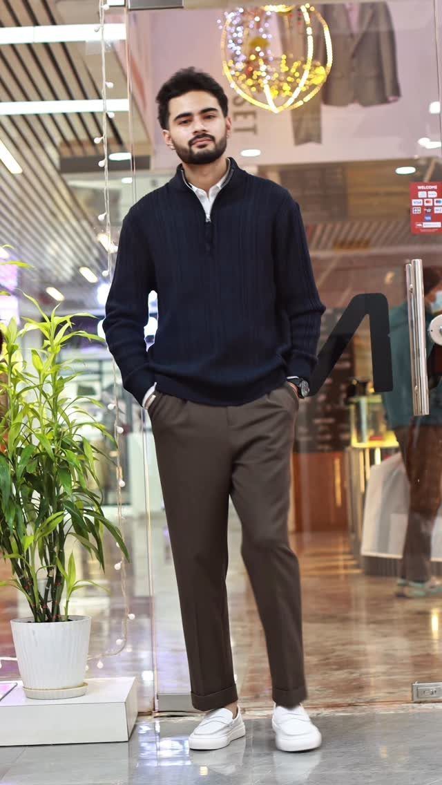 Man wearing a navy cable-knit quarter zip sweater, brown trousers, and white sneakers standing outdoors.