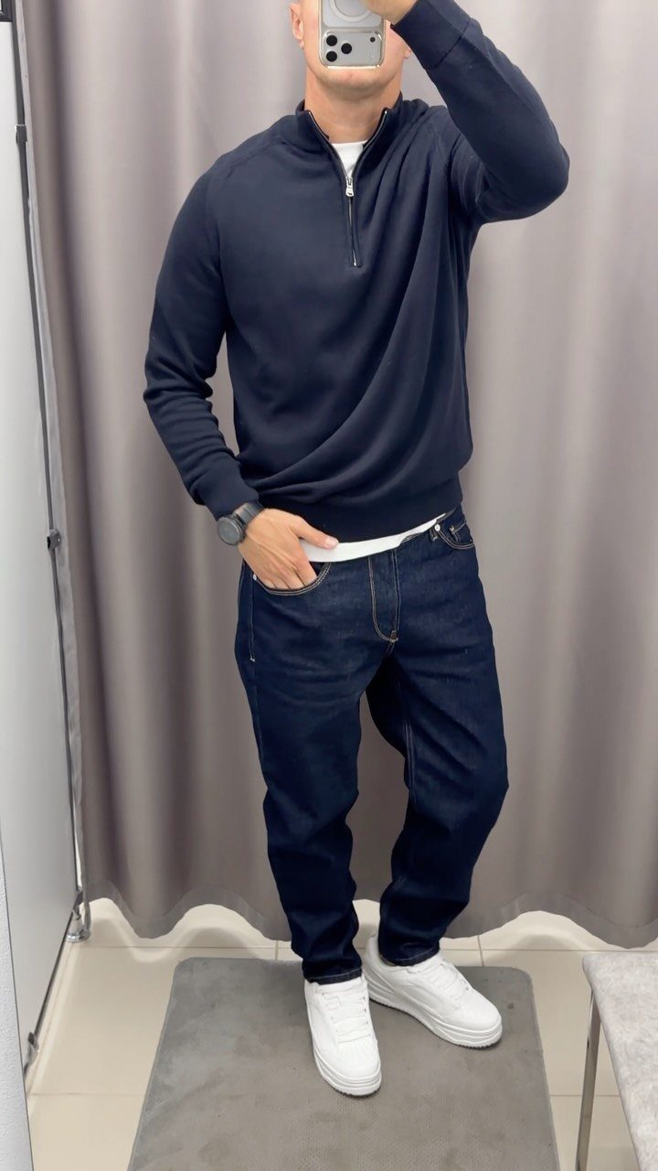 Man wearing a navy quarter-zip pullover, dark jeans, and white sneakers in a fitting room.