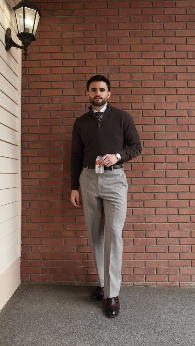 Man posing in a dark brown quarter-zip sweater, white shirt, tie, and grey trousers against a brick wall.