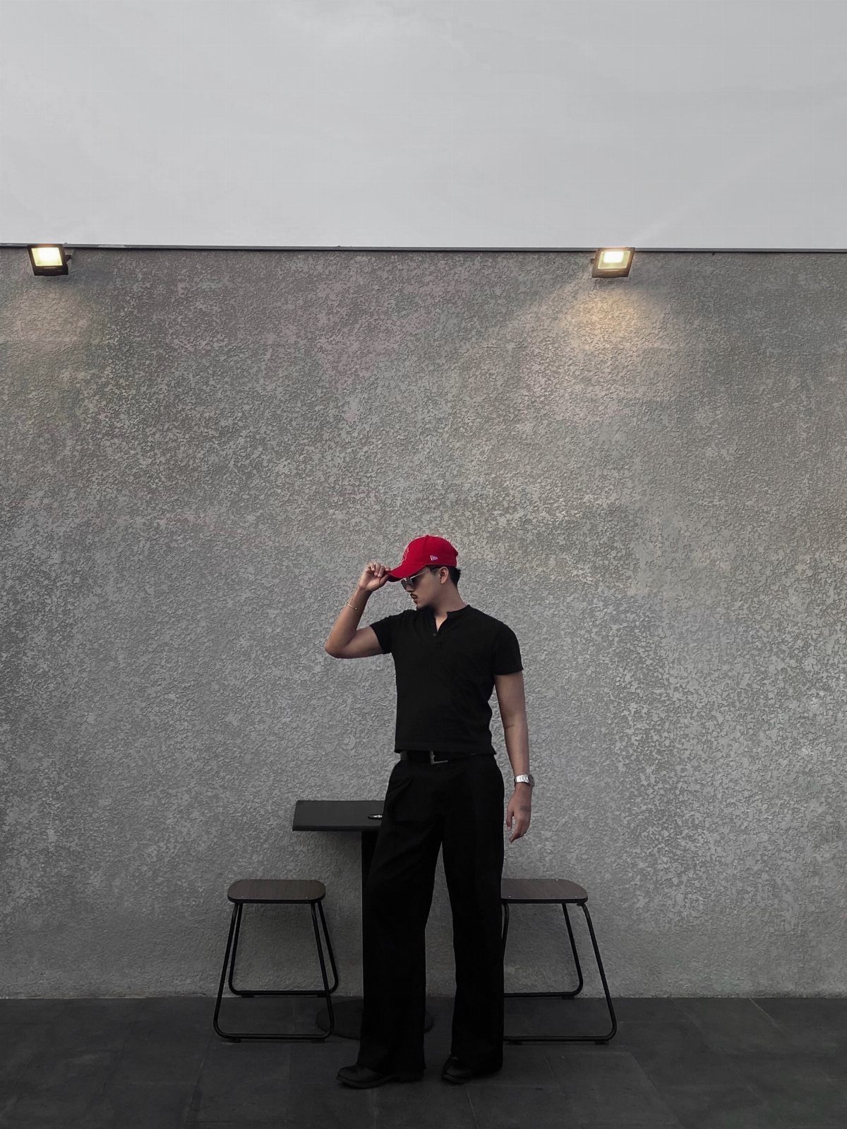 Man in black clothing and a red baseball cap standing against a grey wall.