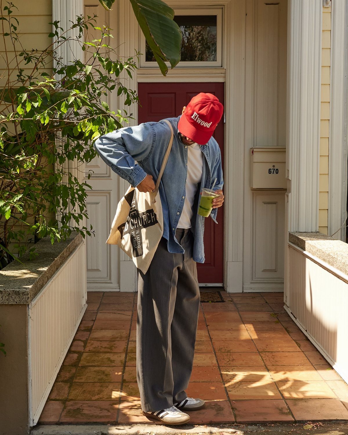 A man in a red hat, denim shirt, and grey trousers standing on a porch.