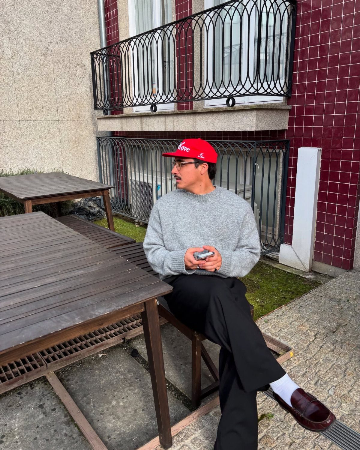 Man in a grey sweater and red hat sitting on a wooden bench outdoors.