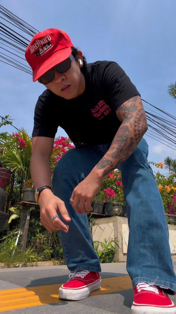 Man in a black t-shirt, blue jeans, red cap, and red sneakers posing outdoors.
