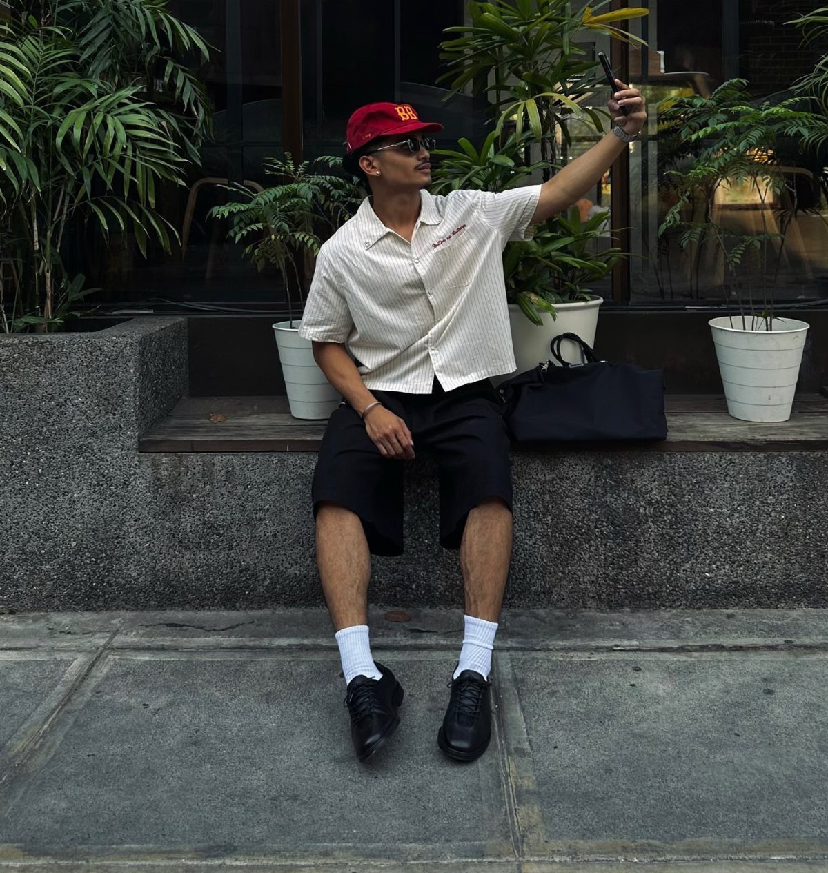 Man in a red cap and patterned shirt sitting on a ledge taking a selfie.