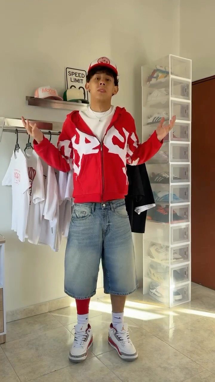 A young man posing in a red patterned hoodie, red hat, denim shorts, and white sneakers.