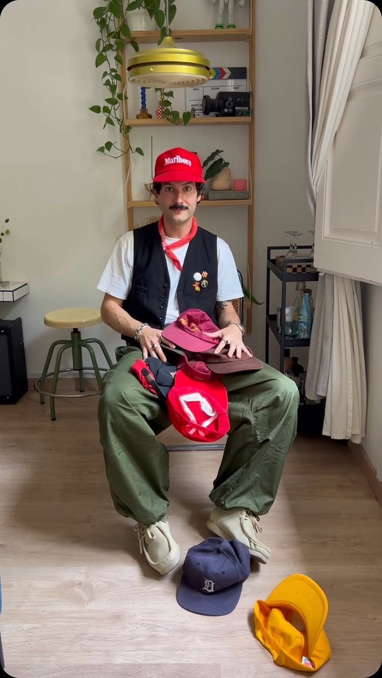 Man wearing a red cap, black vest, and green trousers sitting with various hats.