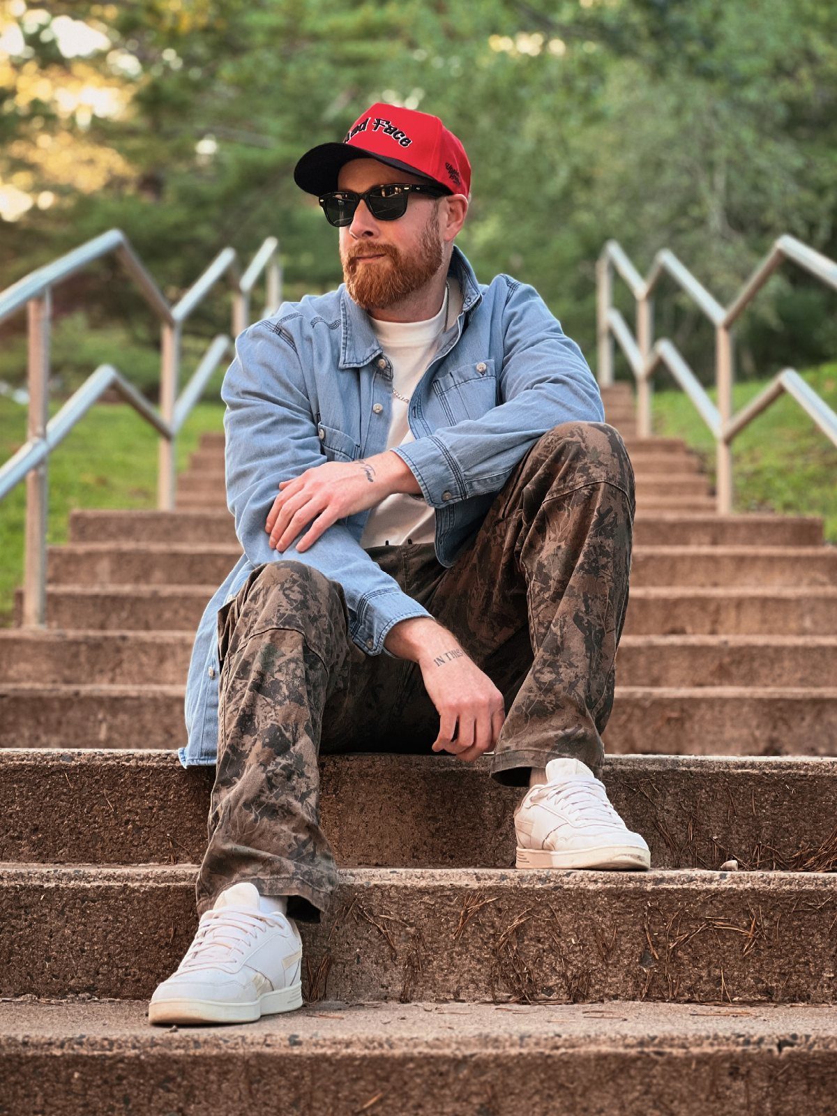 Man in a red cap and denim jacket sitting on outdoor concrete stairs.