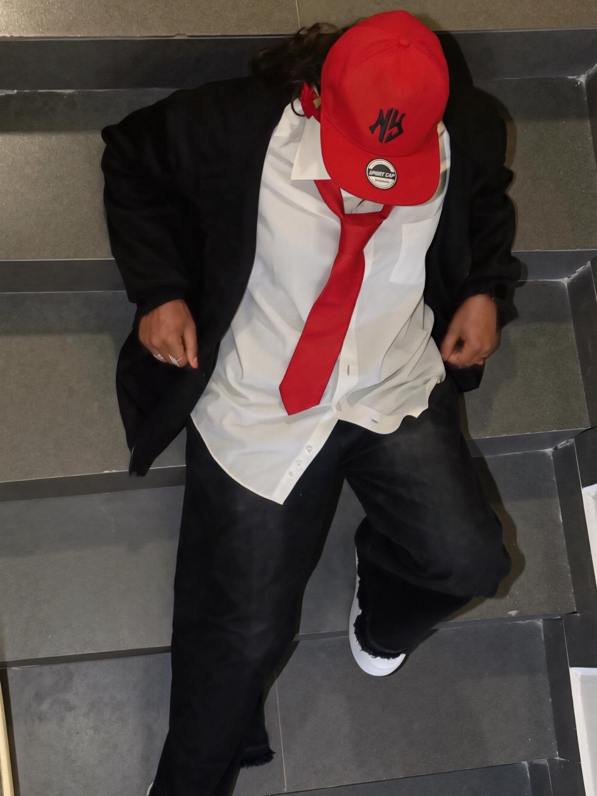 A person in a red hat, red tie, white shirt, and black blazer sitting on stairs.