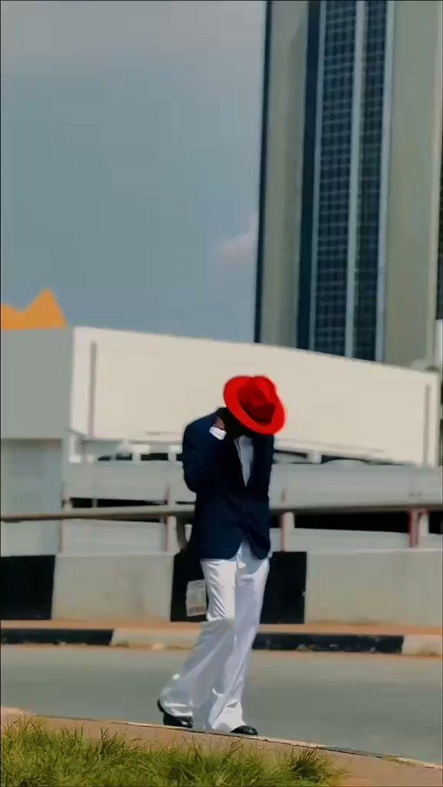 Man in a navy blazer, white pants, and a large red hat walking outdoors.