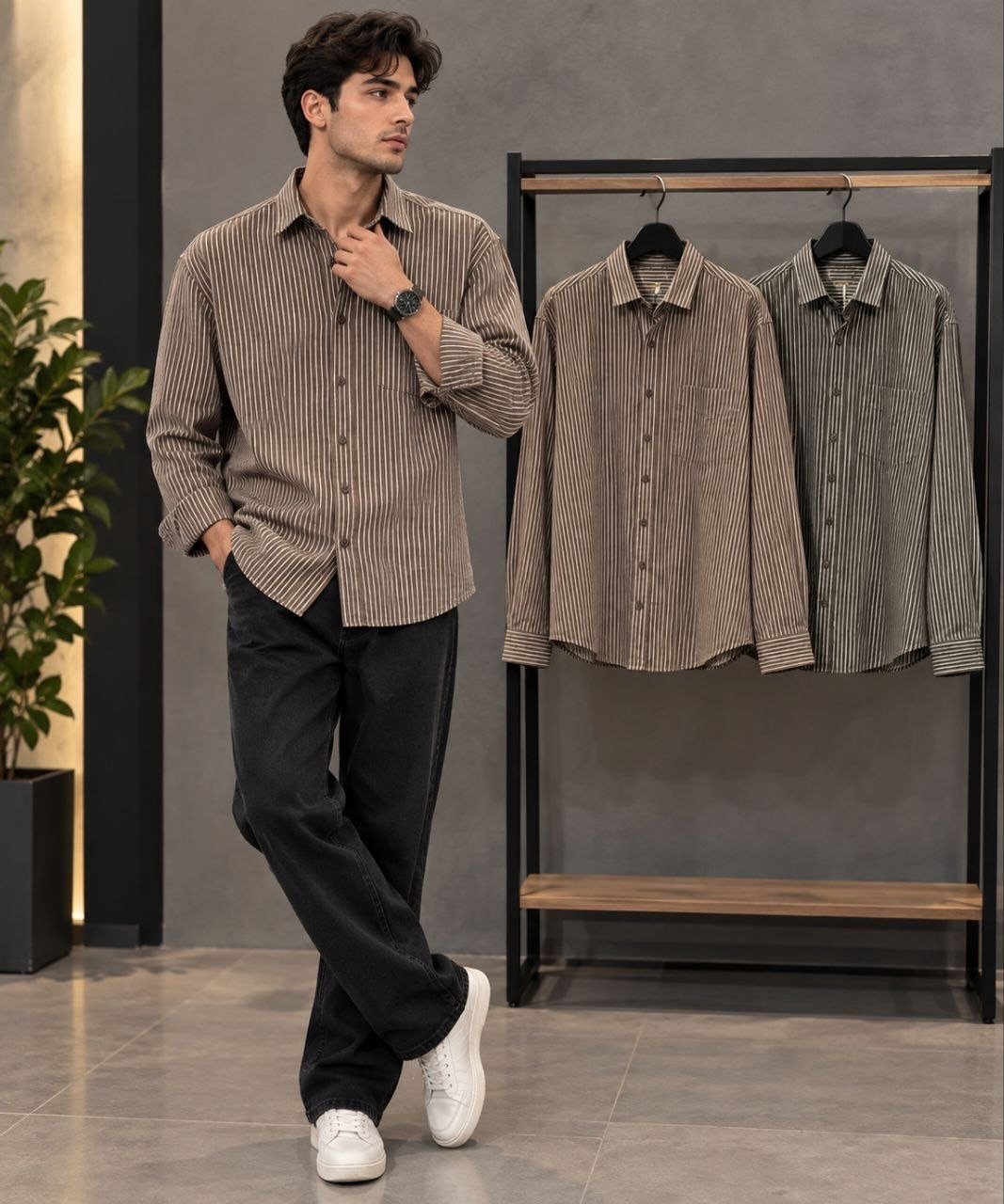 A man posing in a brown striped button-down shirt and black pants next to a clothing rack.