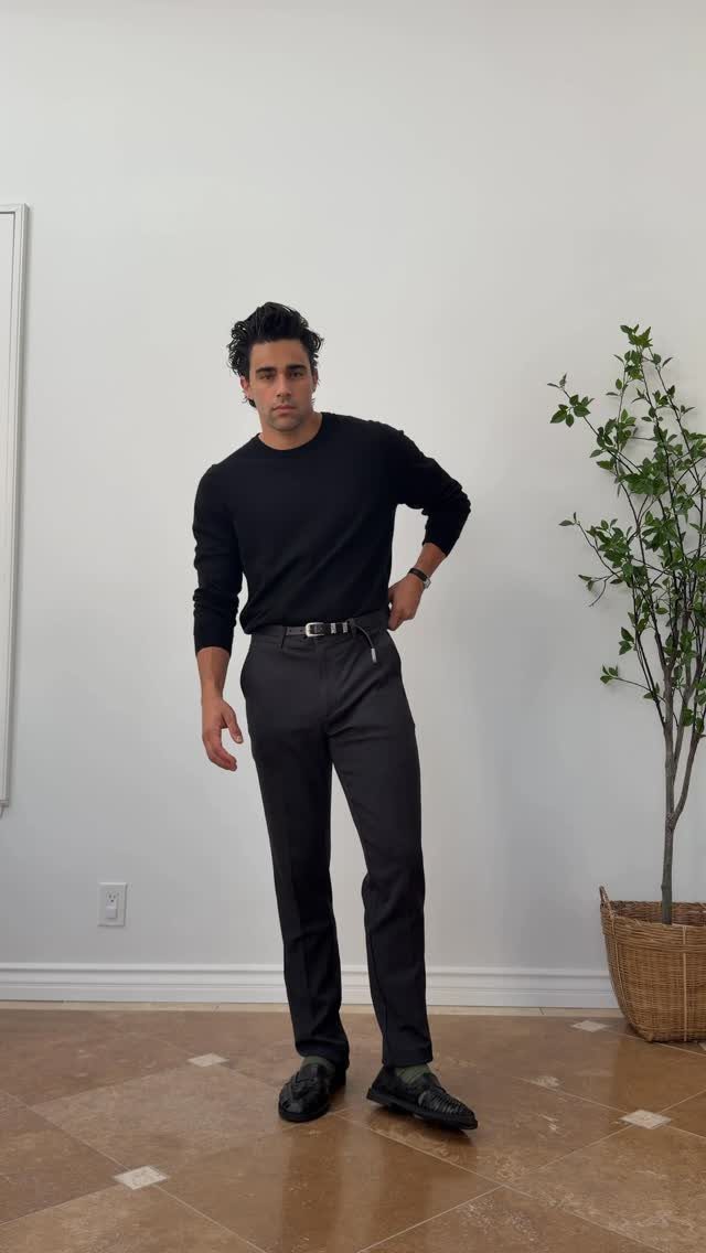 A man posing in a black long-sleeved sweater, dark trousers, and black loafers.