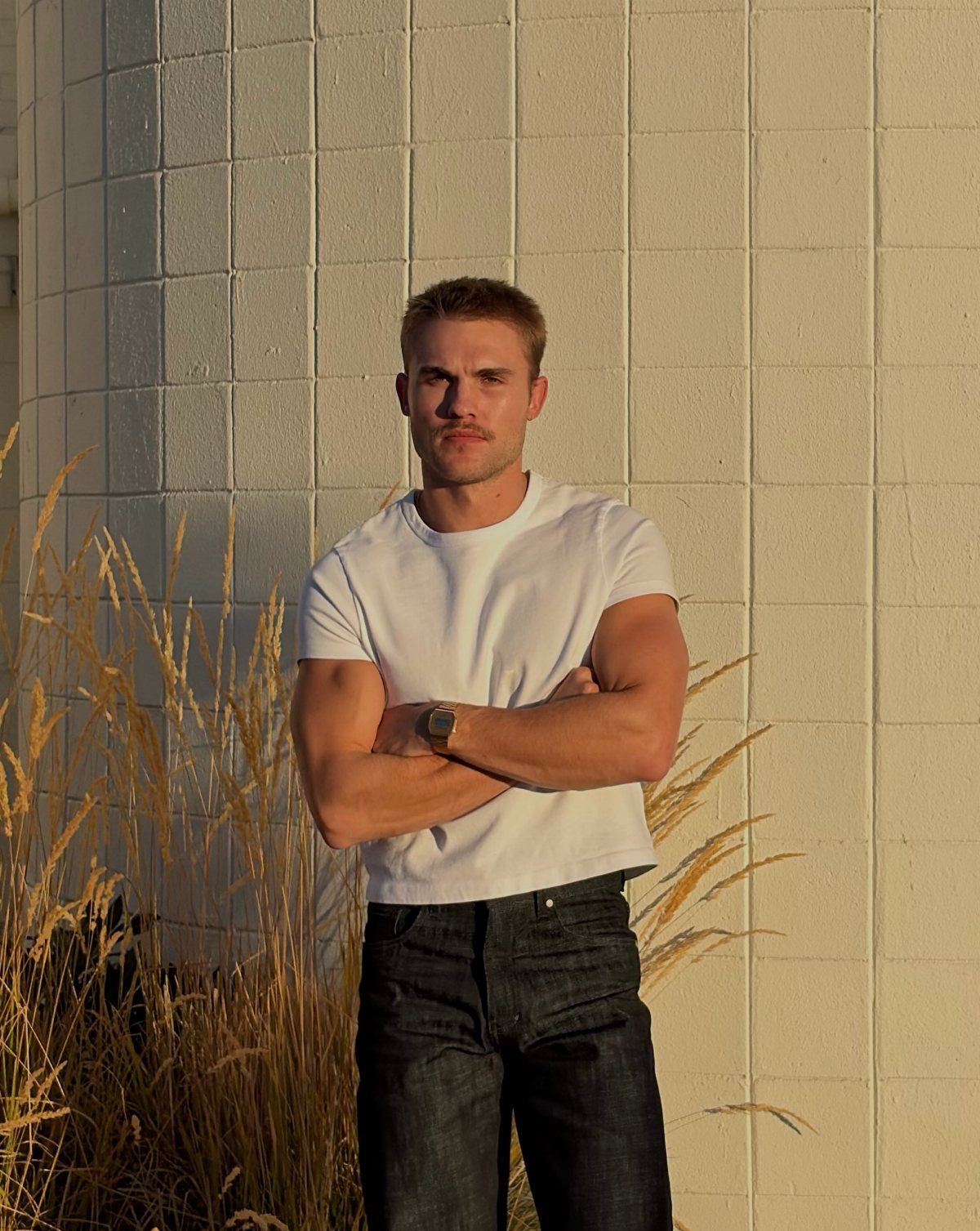 A man in a white t-shirt and dark jeans posing against a white wall.