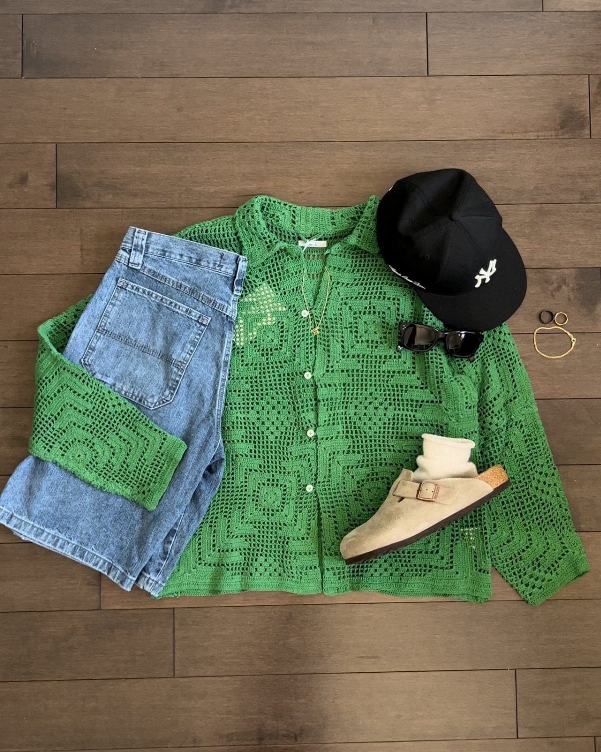 Green crochet shirt, denim shorts, black cap, and tan clogs arranged on a wooden floor.
