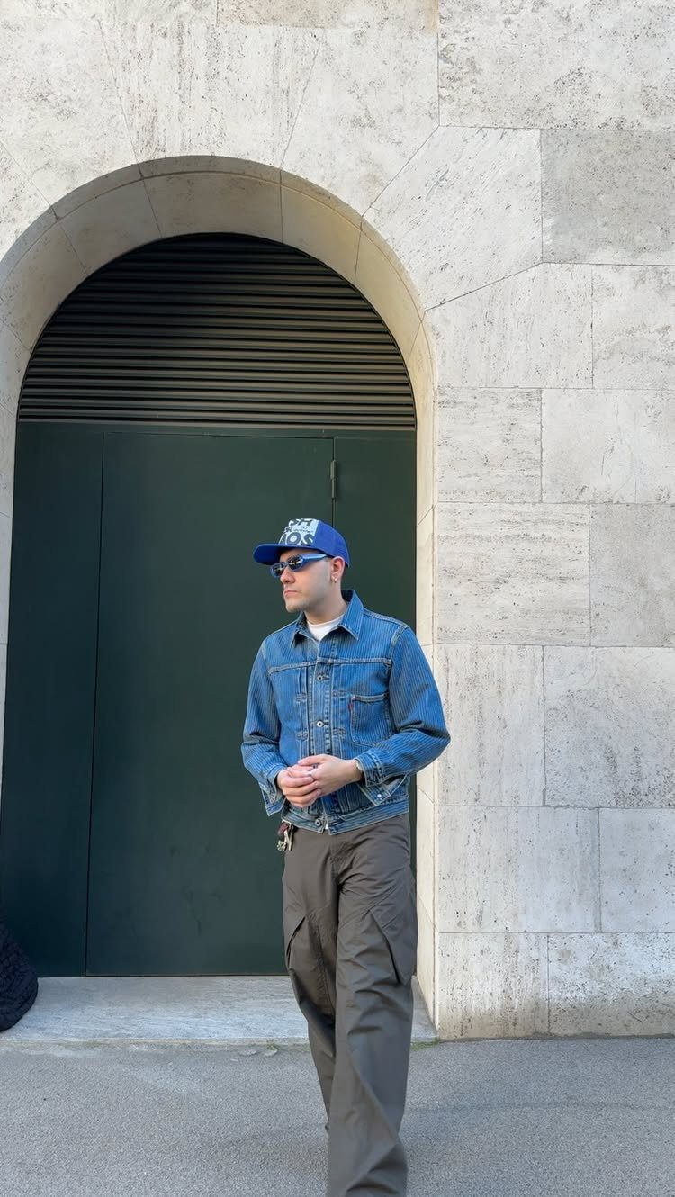 Man wearing a denim jacket, blue cap, sunglasses, and olive cargo pants standing outdoors.