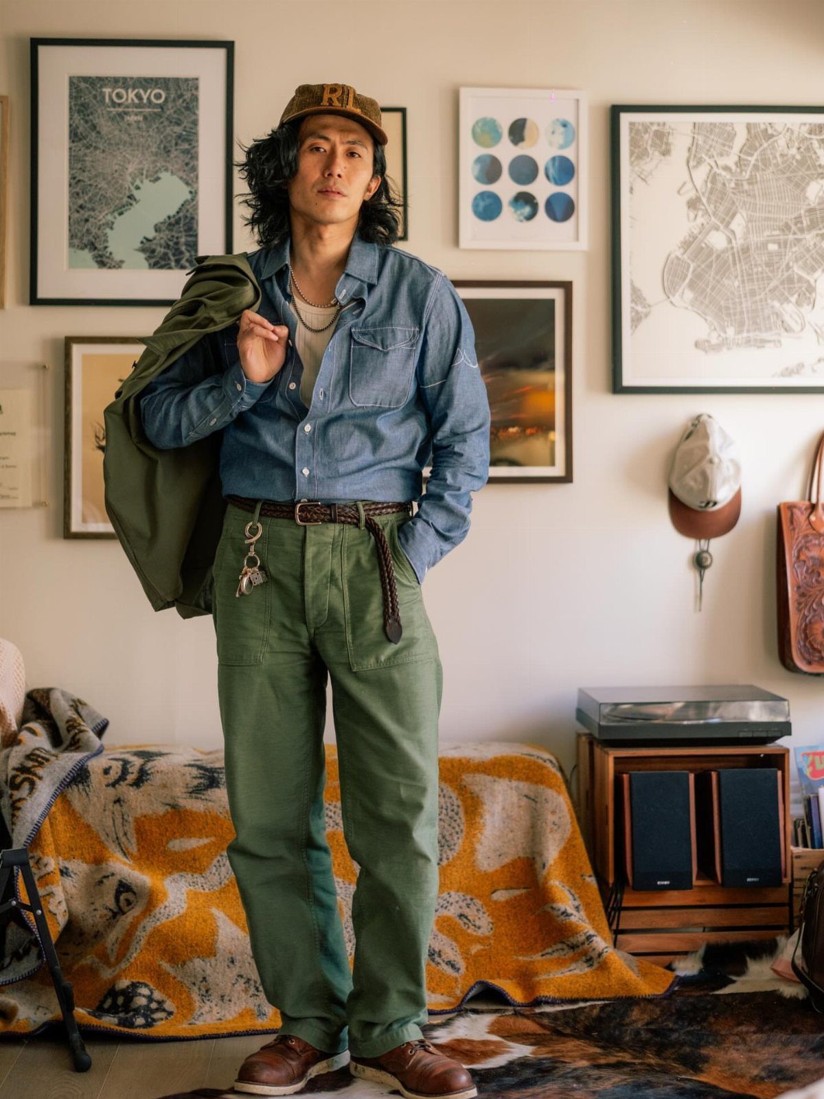 Man standing in a room wearing a denim shirt and green trousers with a jacket over his shoulder.